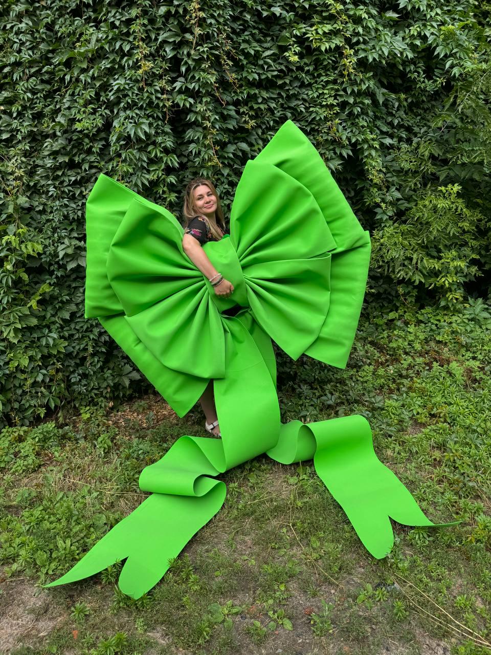May include: An oversized, bright lime green decorative bow constructed from stiff fabric. The bow features two large, pleated loops and two long, trailing ribbon tails that extend downward, creating a substantial and voluminous sculptural form.