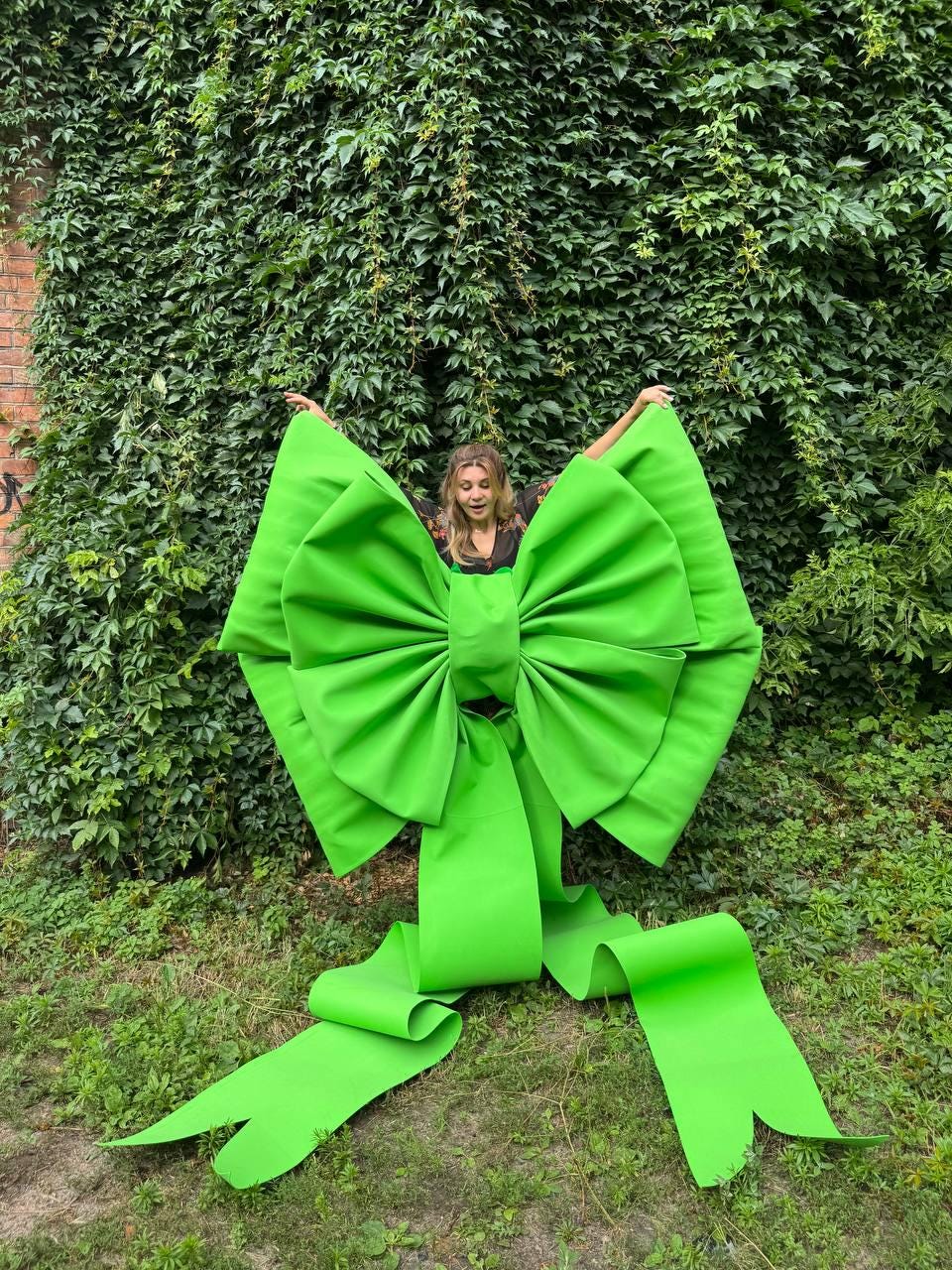May include: An oversized, bright lime green decorative bow featuring two large, pleated loops and two long, trailing ribbon tails that extend downward. The bow is constructed from a smooth, solid-colored material with a structured, symmetrical shape.
