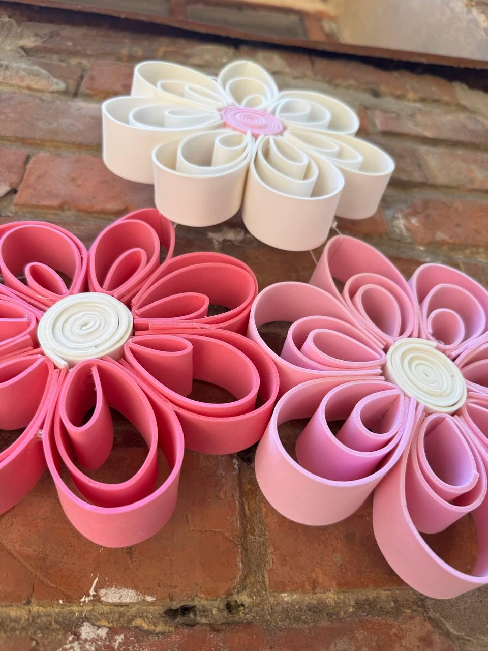 May include: Three flower-shaped wall decorations made from looped strips of foam. One flower is white with a pink center, one is pink with a white center, and one is a lighter shade of pink with a white center. Each features layered petals.