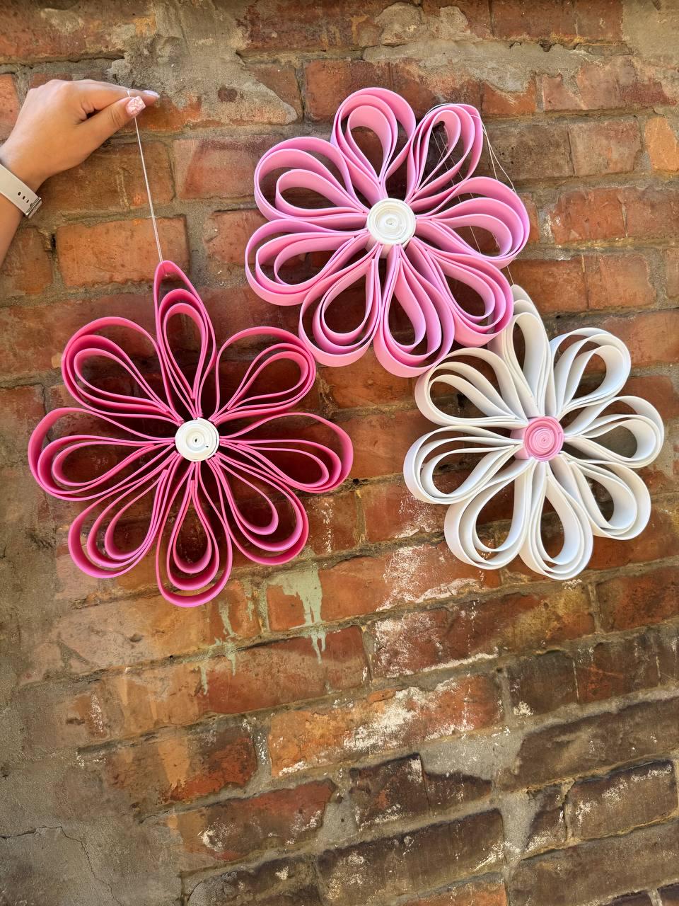 May include: Three decorative flower-shaped wall ornaments constructed from looped paper strips. One flower is dark pink, one is light pink, and one is white. Each flower features a circular center piece and is suspended by a thin white string.