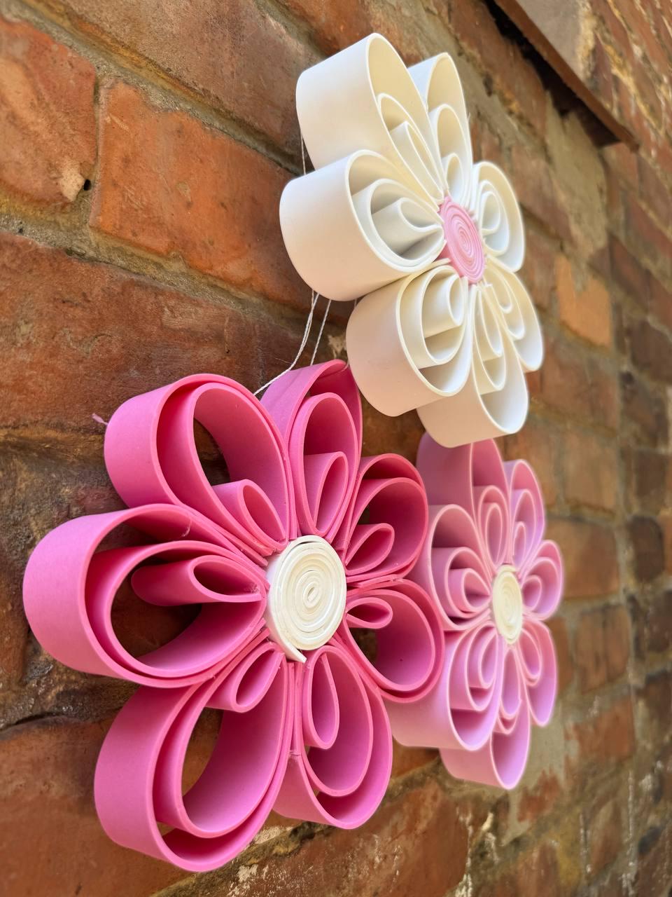 May include: Three dimensional wall art consisting of three flowers crafted from looped strips. One flower is white with a pink center, while the other two are shades of pink with white centers. The petals are formed by folded loops of material.