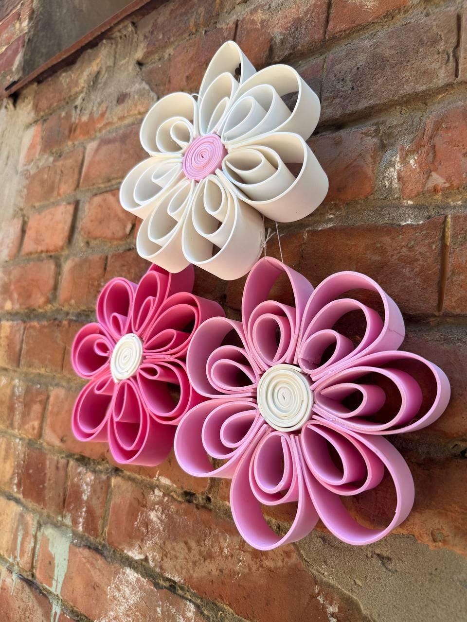 May include: Three decorative wall flowers constructed from looped strips of material. The top flower is white with a pink center, the bottom left is dark pink with a white center, and the bottom right is light pink with a white center.