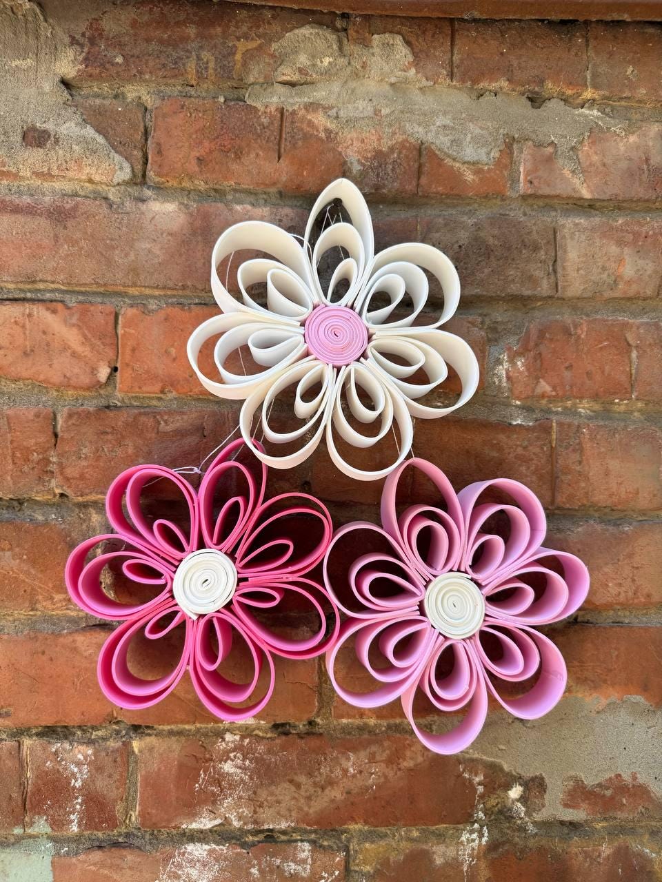 May include: Three decorative flowers crafted from looped strips of material. The top flower is white with a pink center, while the bottom two flowers are shades of pink with white centers. Each flower features six petals formed by folded loops.