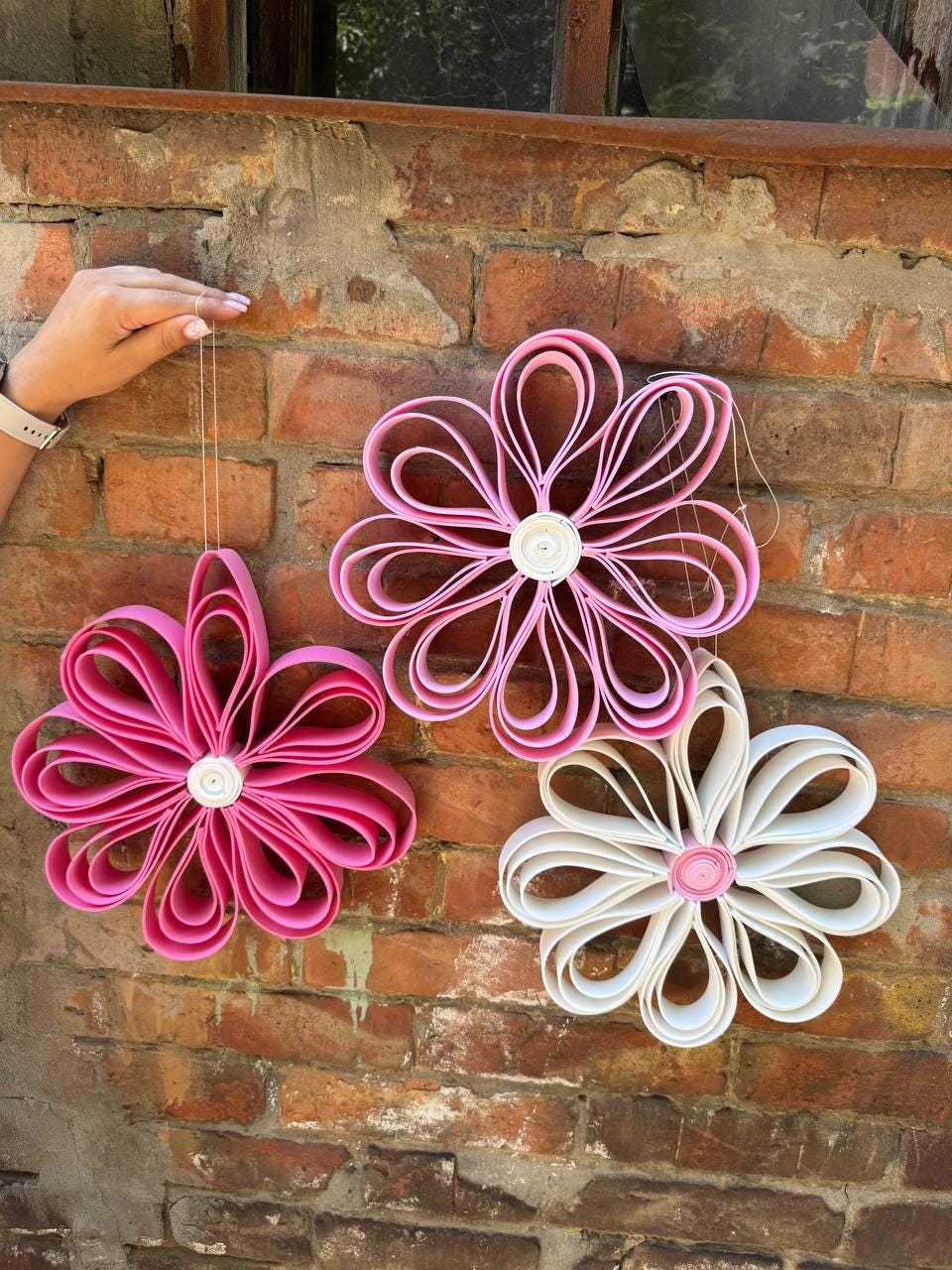 May include: Three hanging flower decorations made from looped strips of paper. One flower is dark pink, one is light pink with a white center, and one is white with a pink center. Each flower features multiple rounded petals arranged in a circular pattern.