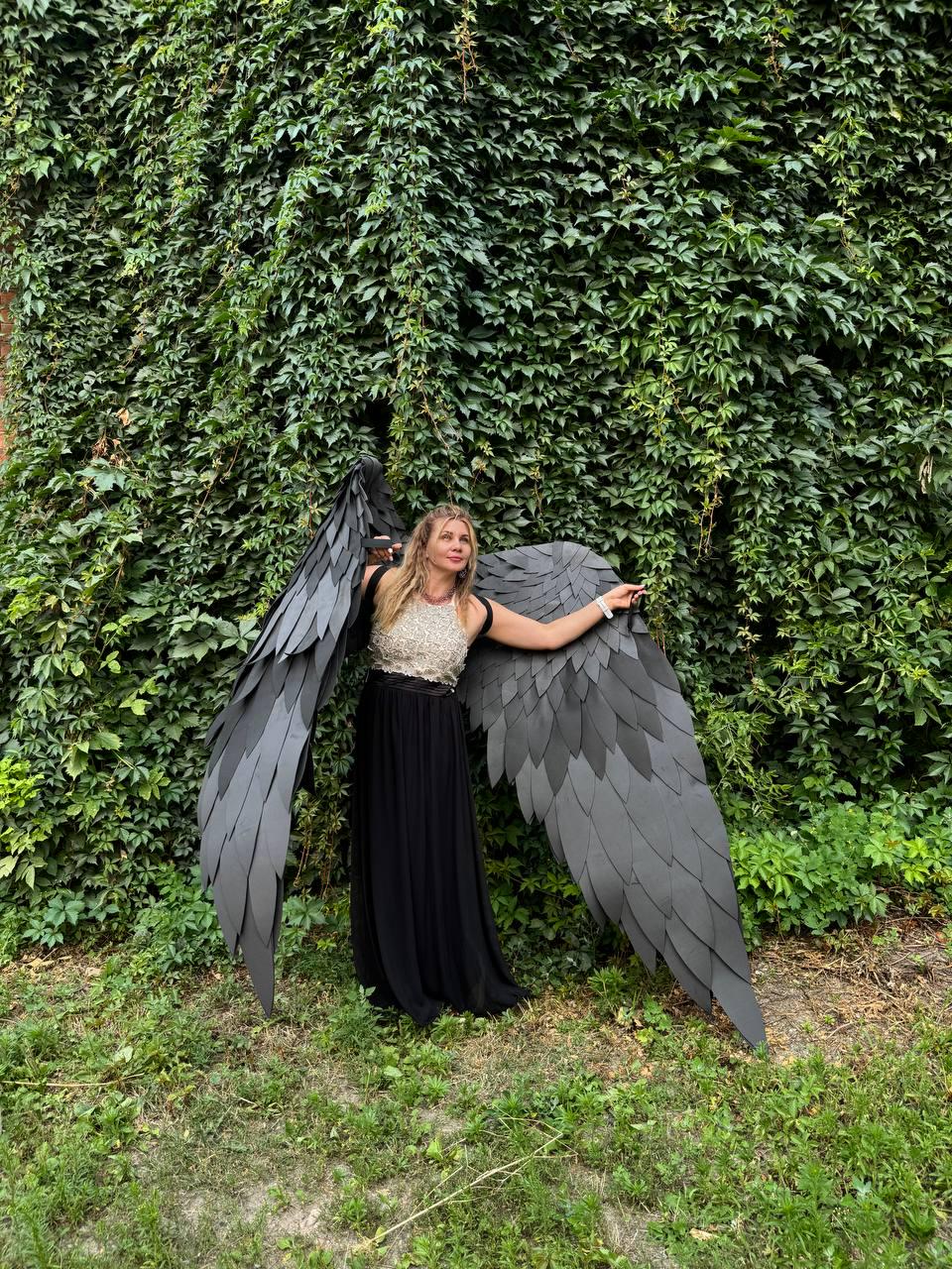 May include: A pair of large, black, layered feather-style wings attached to a sleeveless dress featuring a textured, light-colored bodice and a long, flowing black skirt. The wings extend outward, showcasing a detailed, overlapping petal-like design.