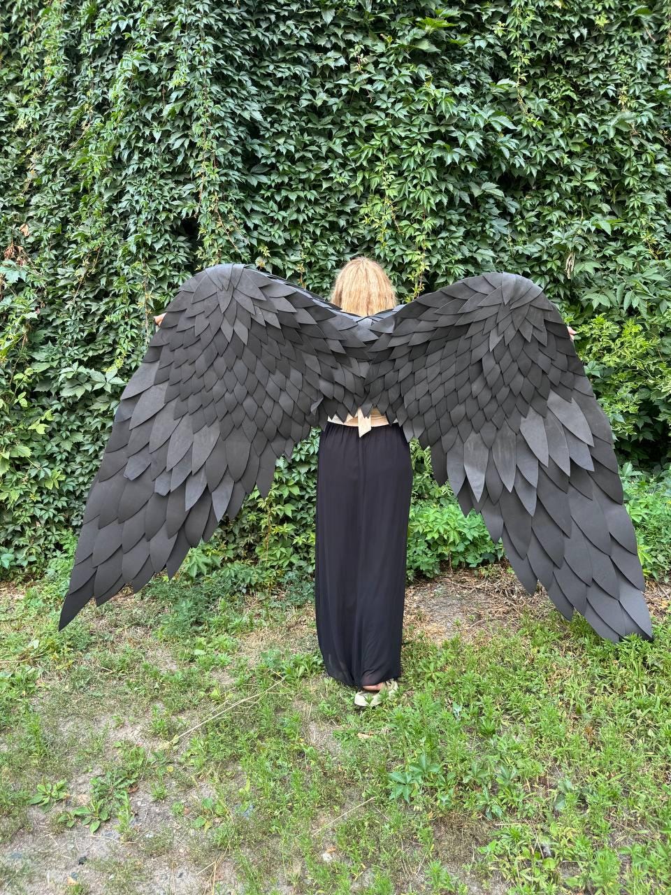 May include: Large, expansive black wings constructed from numerous layered, feather-shaped pieces. The wings are wide and span horizontally, featuring a dark, matte finish with a structured, overlapping design that mimics the appearance of plumage.