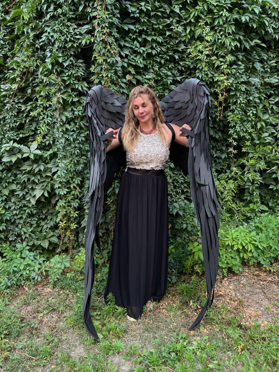 May include: Large black costume wings with layered feather-like panels attached to a harness, worn over a sleeveless top with a metallic textured bodice and a long, flowing black skirt that reaches the ground, creating a dramatic silhouette.