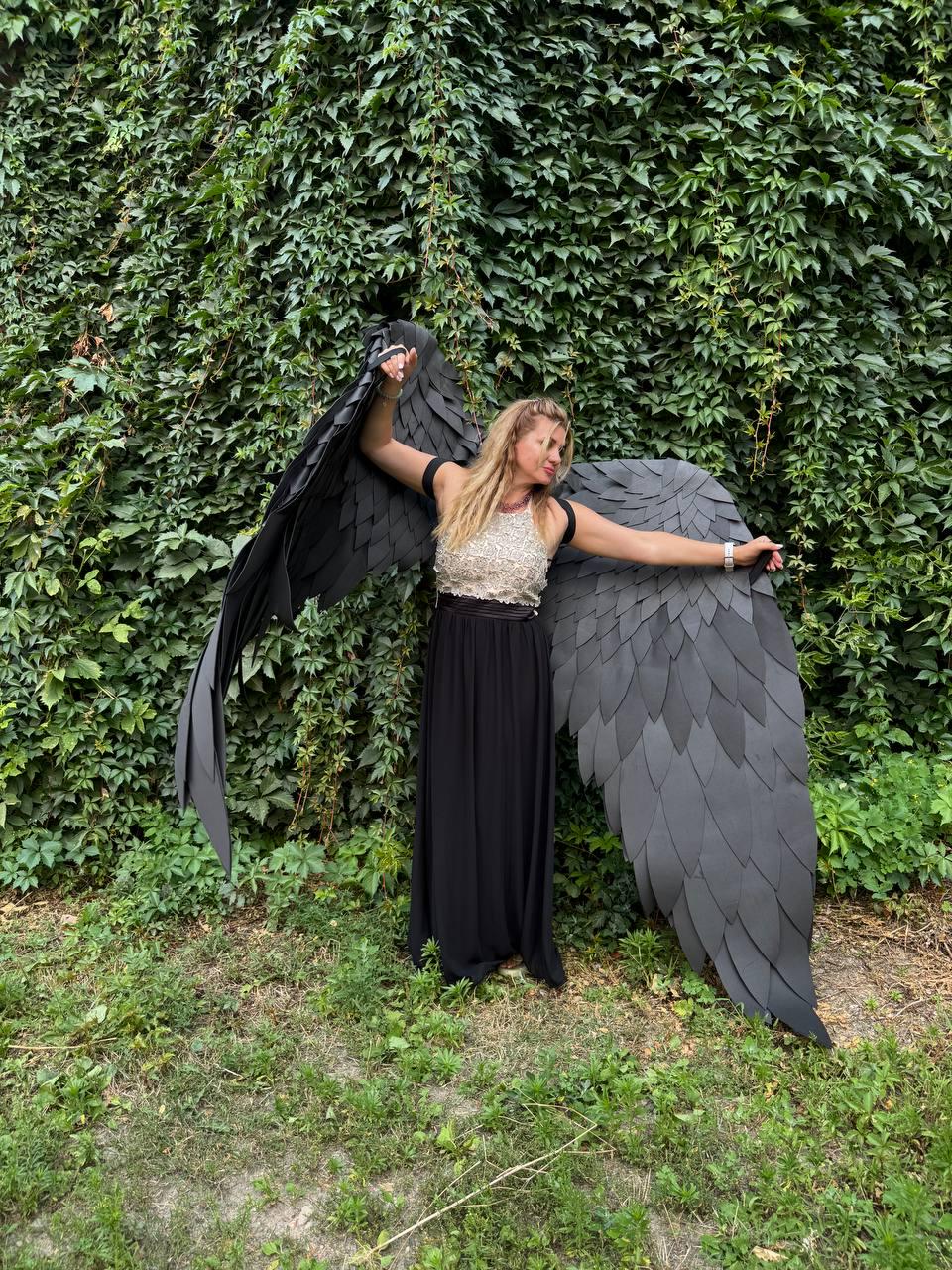 May include: A pair of large, black, articulated wings featuring layered, feather-shaped panels. The wings are worn over a sleeveless, cream-colored, textured bodice paired with a floor-length, solid black skirt, creating a dramatic costume ensemble.