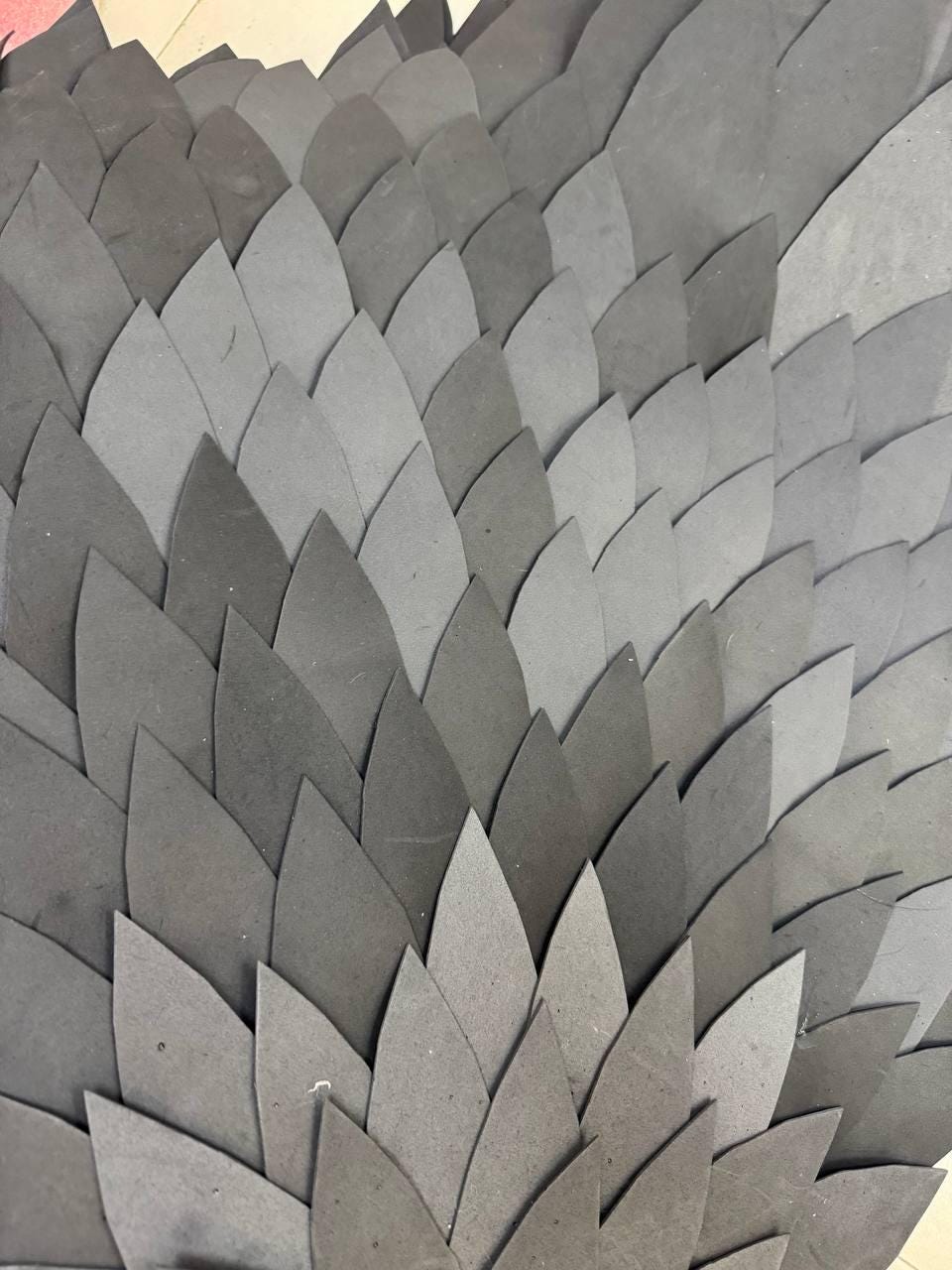 May include: A layered arrangement of numerous dark gray, leaf-shaped cutouts made of a matte, foam-like material. The individual pieces are overlapping in a repeating pattern, creating a textured, scale-like surface with varying shades of gray.