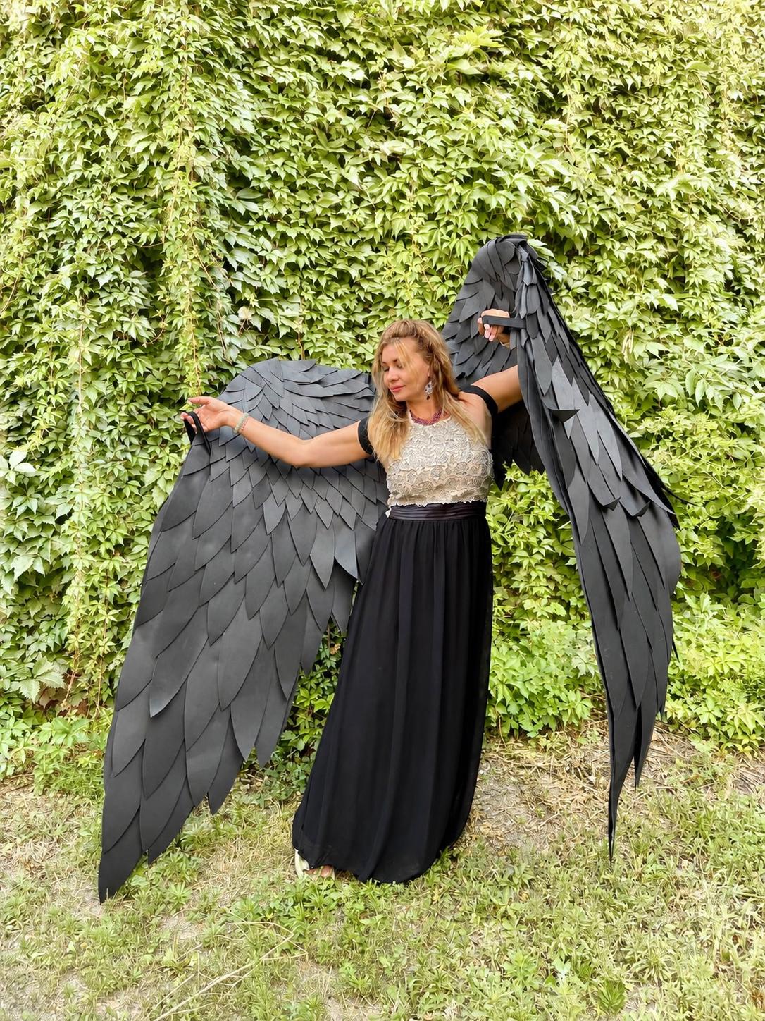 May include: Large black wings constructed from layered, feather-shaped panels attached to a sleeveless dress featuring a textured, light-colored bodice and a long, flowing black skirt that reaches the ground, creating a dramatic silhouette.