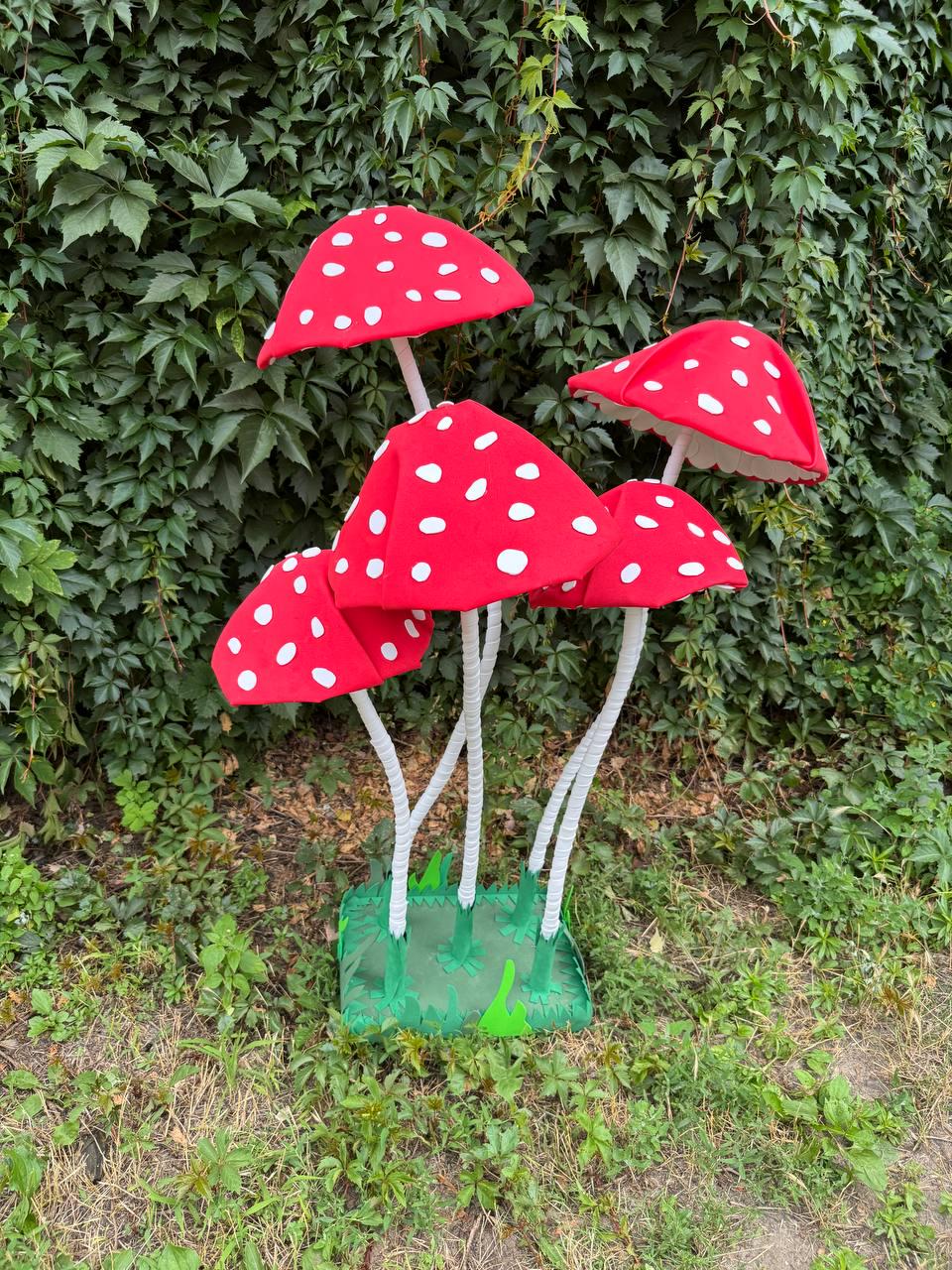 May include: A decorative display featuring five red mushroom caps with white spots, each supported by a thin white stem. The mushrooms are attached to a square green base that features a cutout grass pattern along its edges.