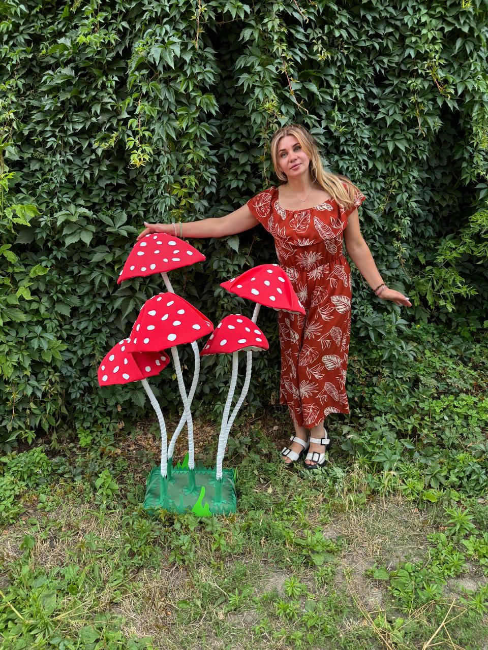May include: A set of four large, decorative artificial mushrooms with bright red caps featuring white polka dots and white stems, mounted on a green base, standing alongside a rust-colored jumpsuit with a white leaf pattern.