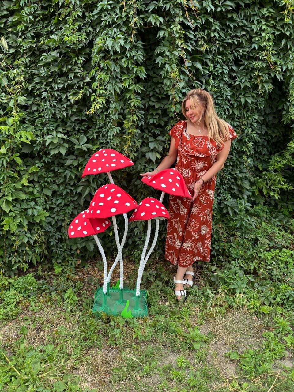 May include: A set of four decorative artificial mushrooms featuring bright red caps with white polka dots and long, slender white stems, all mounted on a green square base, standing alongside a rust-colored jumpsuit with a white leaf pattern.