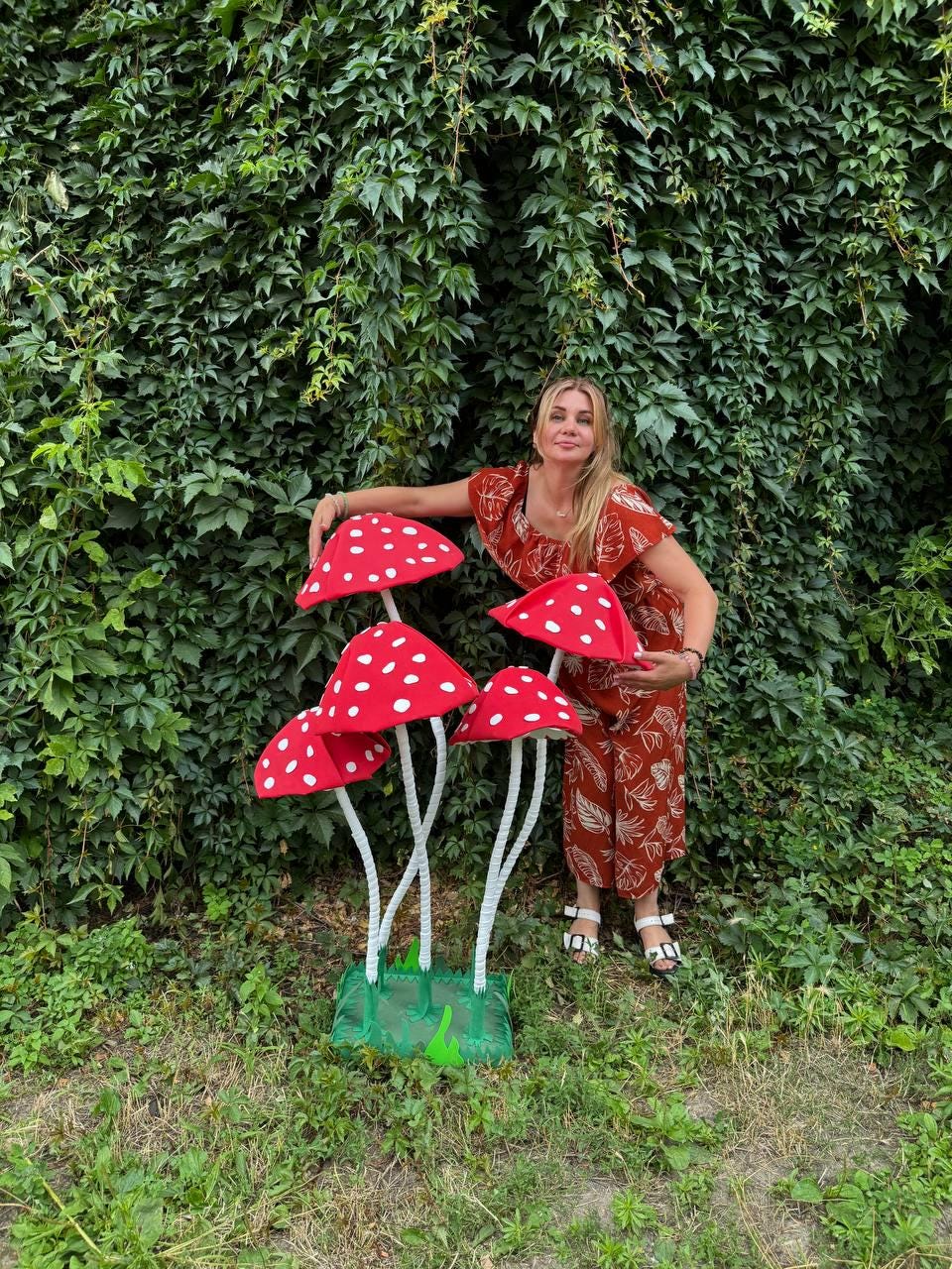 May include: A decorative floor sculpture featuring five red mushroom caps with white spots, supported by thin white stems rising from a green rectangular base. The mushrooms are arranged at varying heights to create a cluster of toadstools.