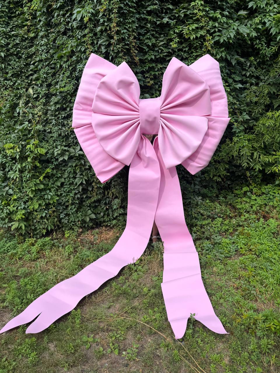 May include: A large, oversized decorative bow in a solid light pink color. The bow features two wide, pleated loops and two long, trailing ribbon tails that extend downward, ending in notched, V-shaped cutouts at the bottom edges.