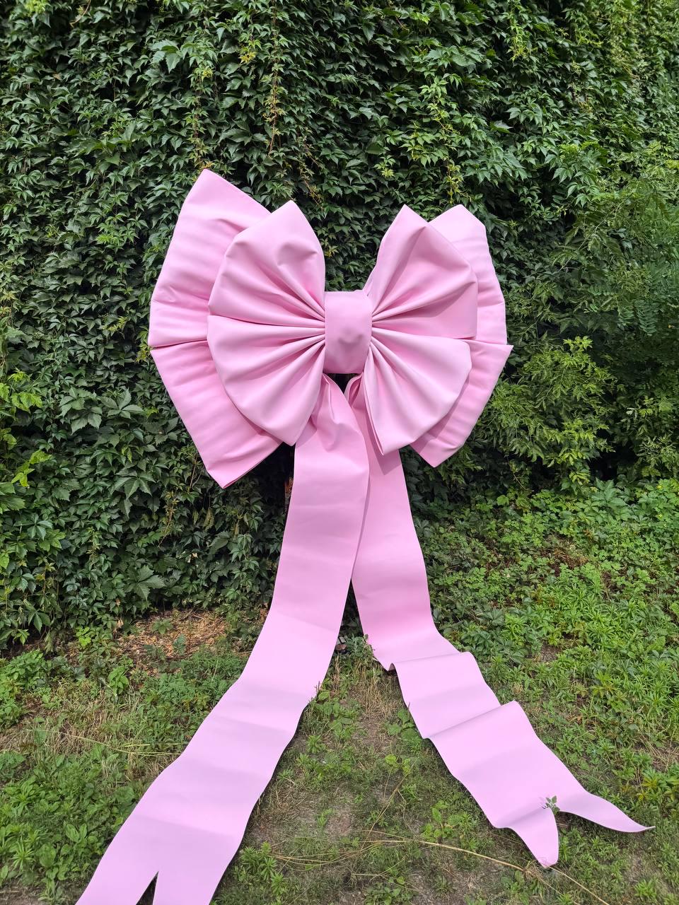 May include: A large, light pink decorative bow made of smooth material, featuring two wide, pleated loops and two long, trailing ribbon tails that extend downward, creating a symmetrical and structured appearance with soft, matte-finish surfaces.