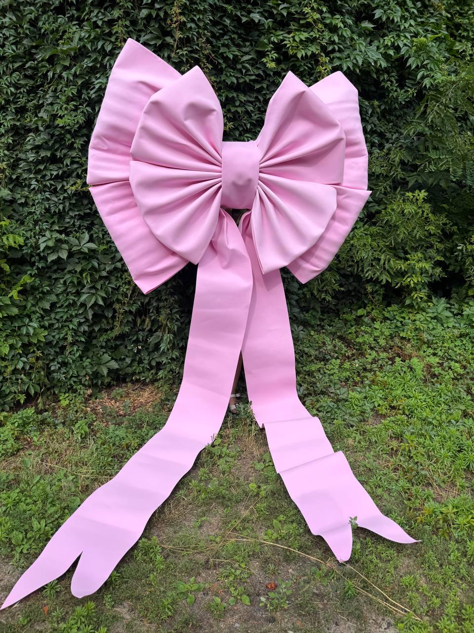 May include: A large, light pink decorative bow featuring a central knot and two long, flowing ribbon tails that extend downward. The bow is constructed with structured, smooth material and displays prominent folds and pleats throughout its design.