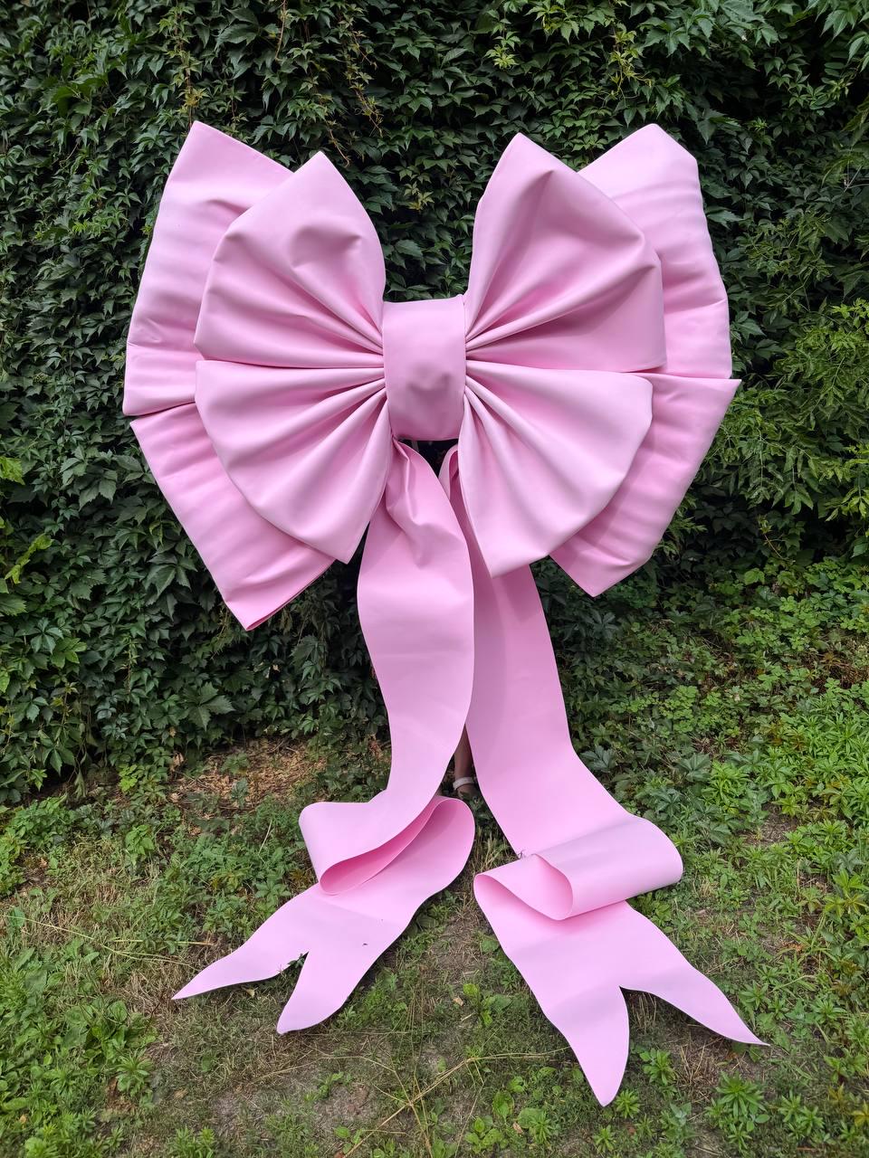 May include: A large, light pink decorative bow featuring a central knot and two long, flowing ribbon tails that extend downward. The bow is constructed with multiple folds and pleats, creating a voluminous, structured appearance with a smooth finish.