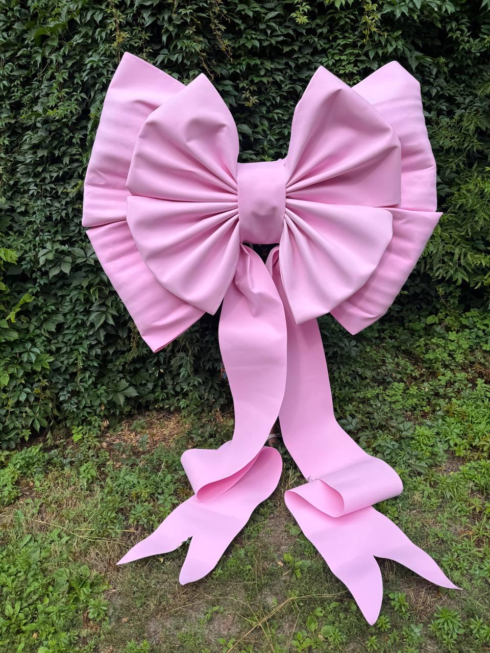 May include: A large, light pink decorative bow featuring a central knot and two wide, pleated loops on each side. Two long, flowing ribbons extend downward from the center, each ending in a V-shaped notch at the bottom edge of the fabric.