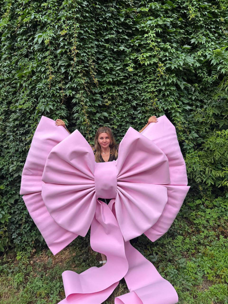 May include: An oversized, light pink decorative bow constructed from smooth, matte material. The bow features two large, voluminous loops and two long, wide ribbon tails that extend downward, creating a prominent and structured appearance.