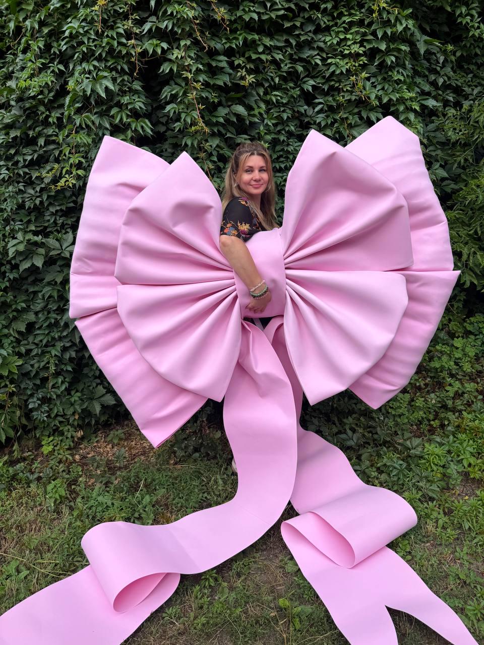 May include: An oversized, light pink decorative bow constructed from smooth, matte material. The bow features two large, voluminous loops and two long, trailing ribbon tails that extend downward, creating a substantial and prominent display piece.