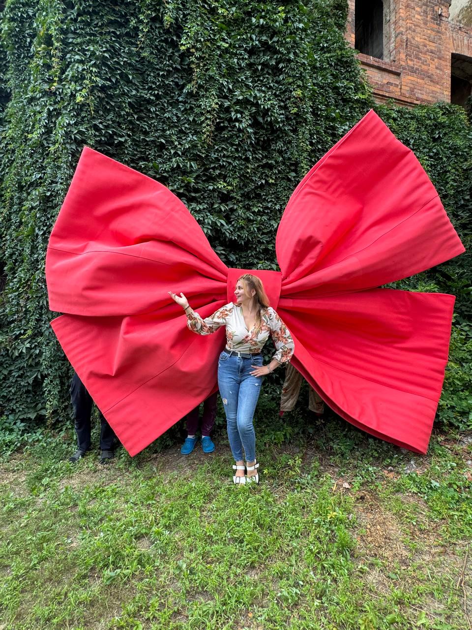 May include: A giant, oversized red fabric bow with prominent folds and a central knot. The bow is constructed from a smooth, matte red material and is positioned upright, displaying large, symmetrical wings that extend horizontally across the frame.