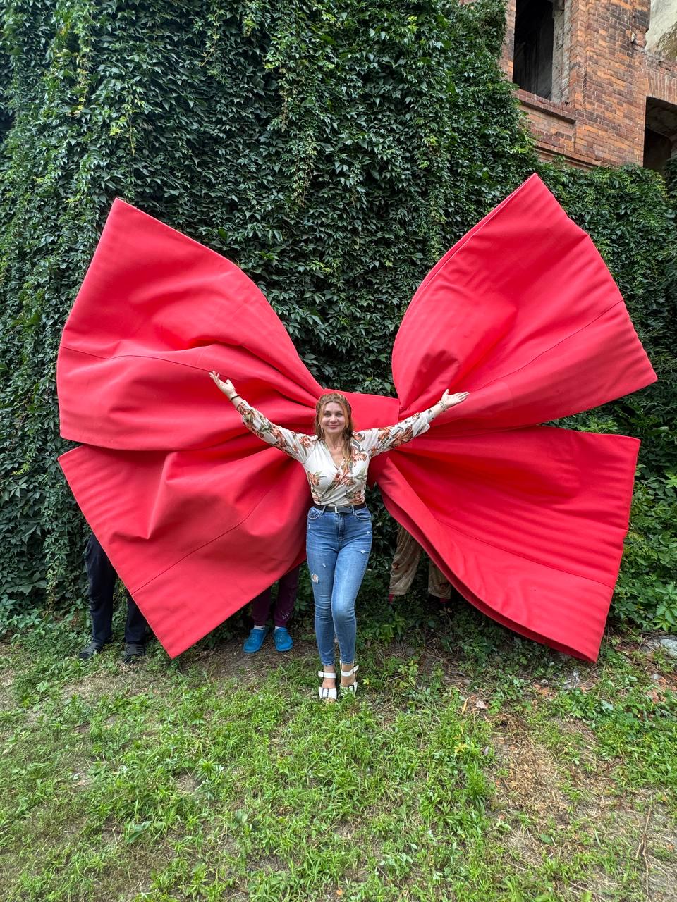 May include: A giant, oversized red fabric bow with wide, structured loops and long, flowing tails. The bow is constructed from a smooth, solid red material and is positioned behind a person wearing a floral long-sleeve top and blue jeans.