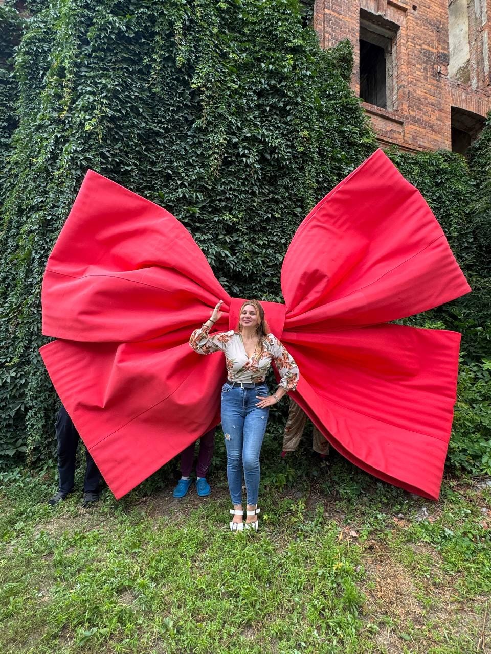 May include: A massive, oversized red fabric bow featuring large, structured loops and long, flowing tails. The bow is constructed from a vibrant, solid red material with visible seams and folds, creating a dramatic, expansive display piece.