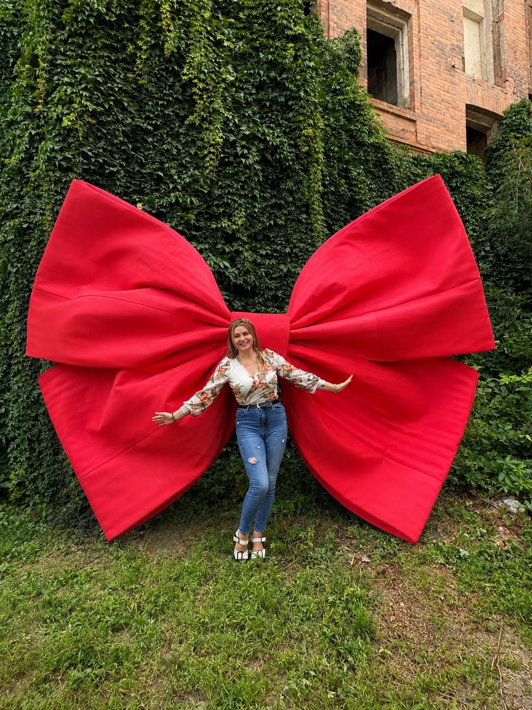 May include: A large, bright red decorative bow made of fabric, featuring wide, structured loops and a central knot. The bow is positioned against a wall covered in dense green ivy, creating a striking contrast between the vibrant red and the foliage.