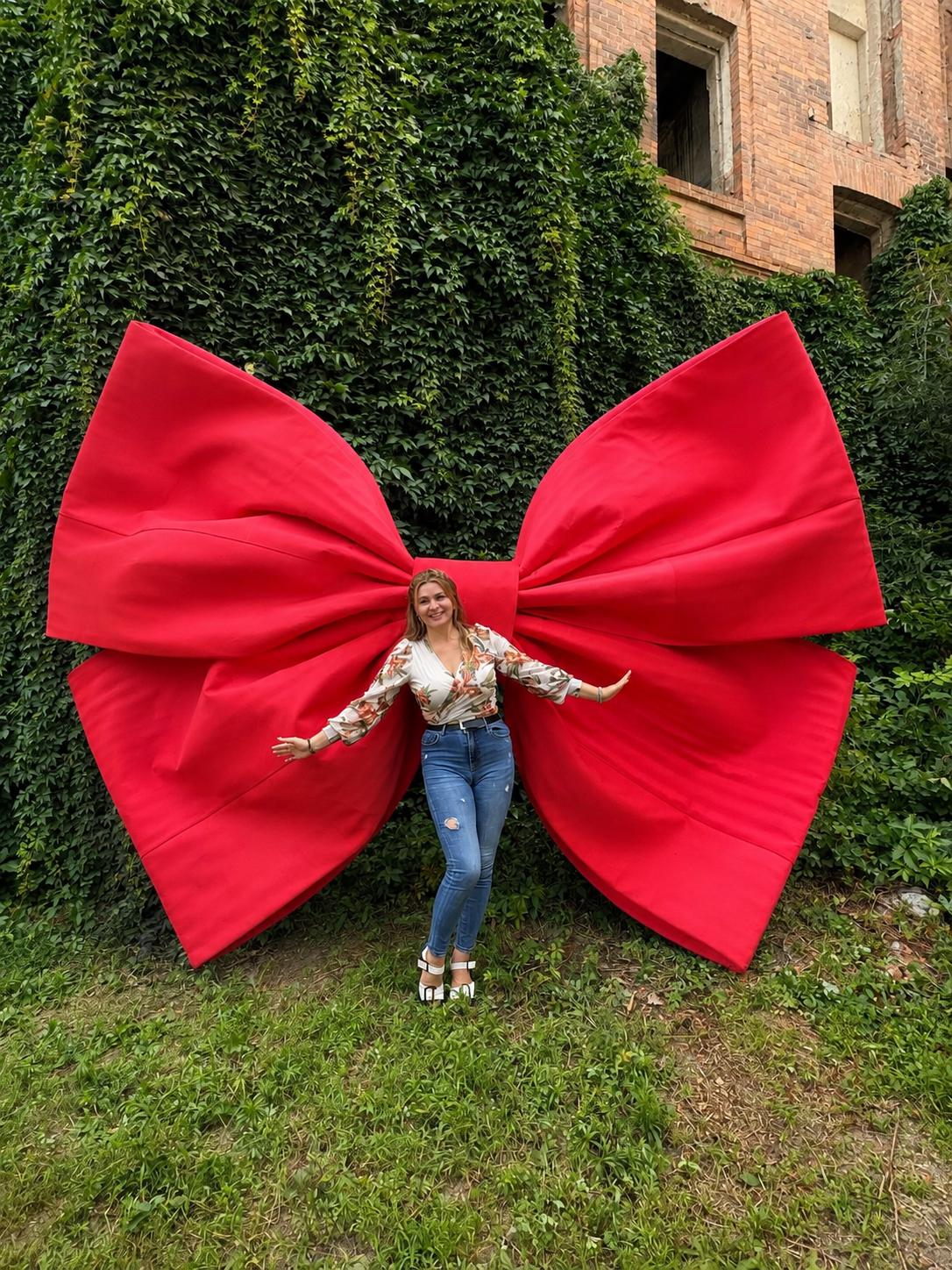 May include: A large, bright red decorative bow made of fabric, featuring wide, structured loops and a central knot. The bow is positioned against a wall covered in dense green ivy, creating a striking contrast between the vibrant red and the foliage.