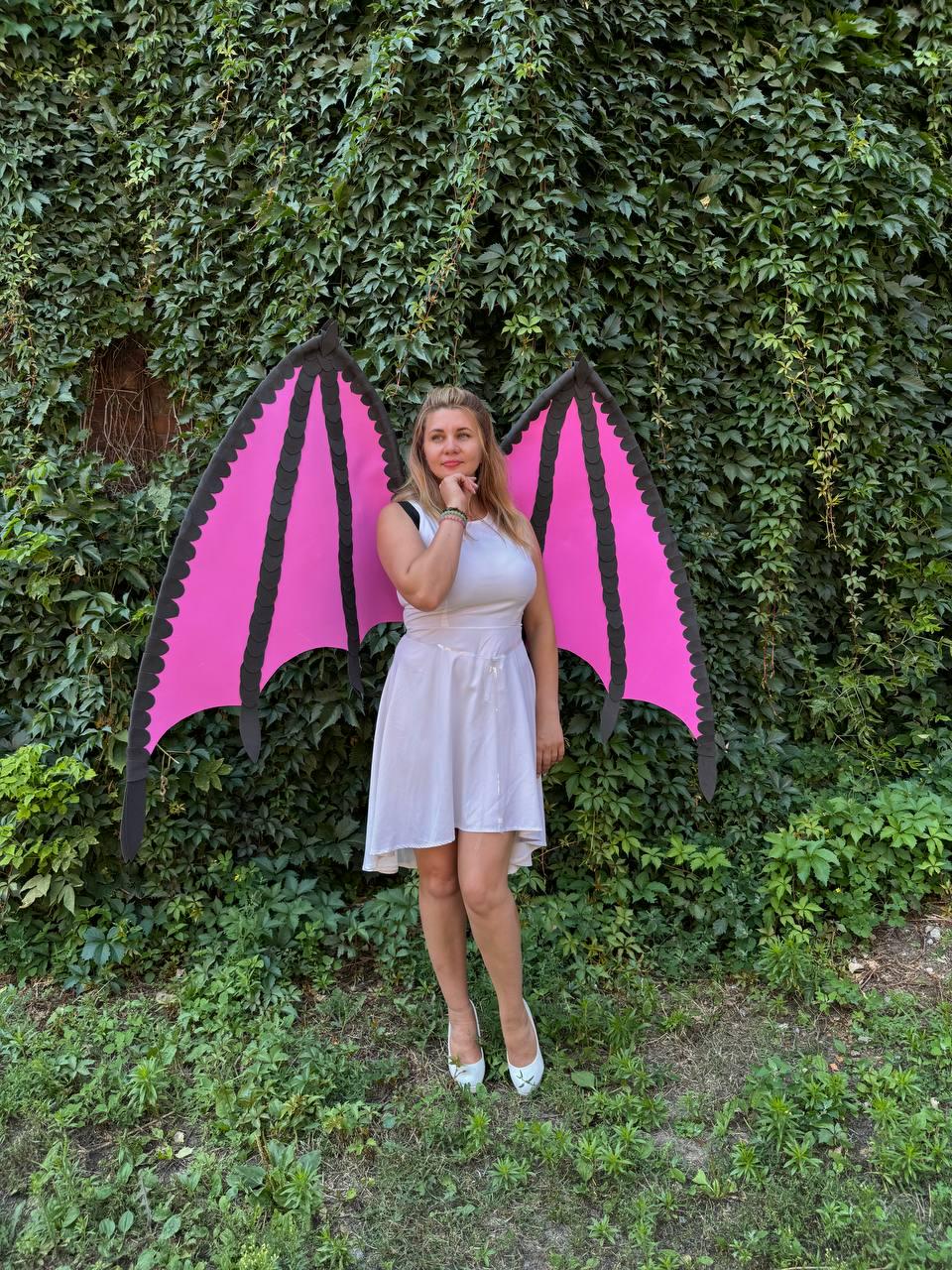 May include: A white sleeveless dress paired with large, detachable bat-like wings. The wings feature a vibrant pink interior with black scalloped edges and black structural ribs, extending outward from the back of the white dress.