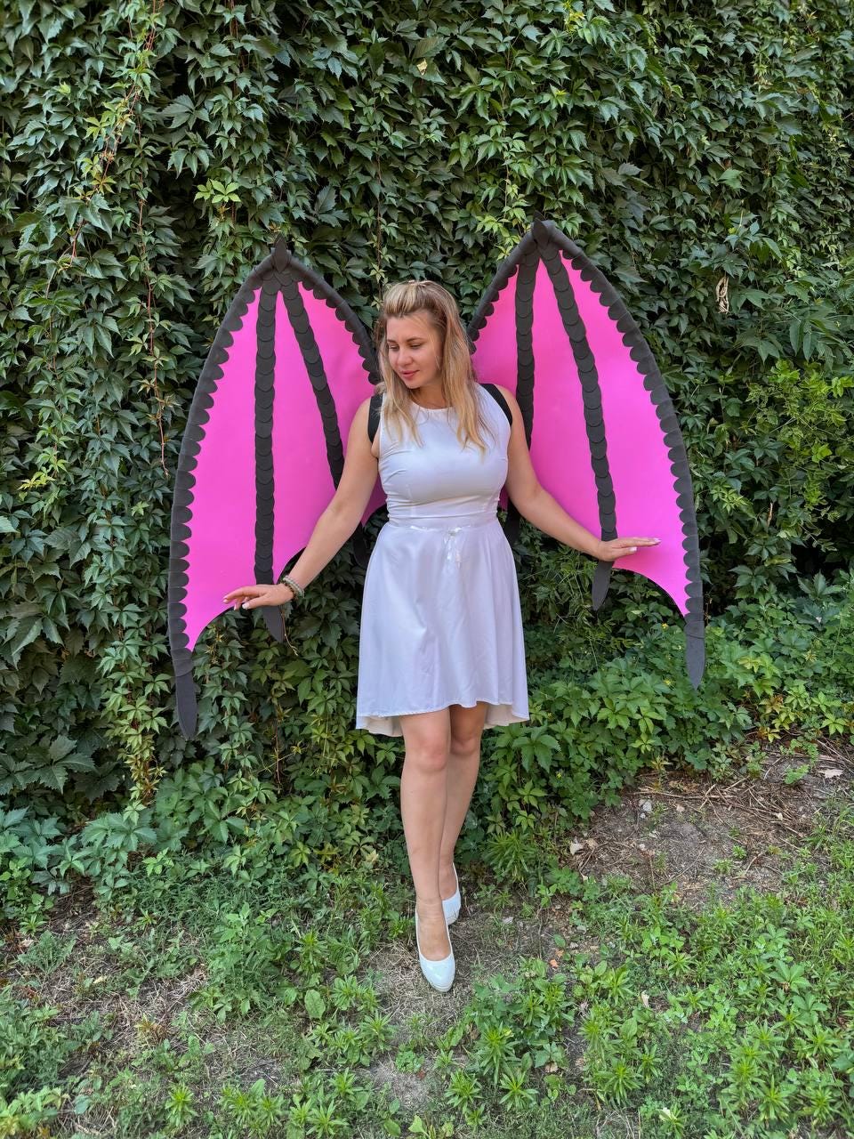 May include: A white sleeveless dress with a high-low hemline paired with large, detachable dragon-style wings. The wings feature bright pink panels framed by thick black borders with scalloped edges, secured by black straps worn over the shoulders.