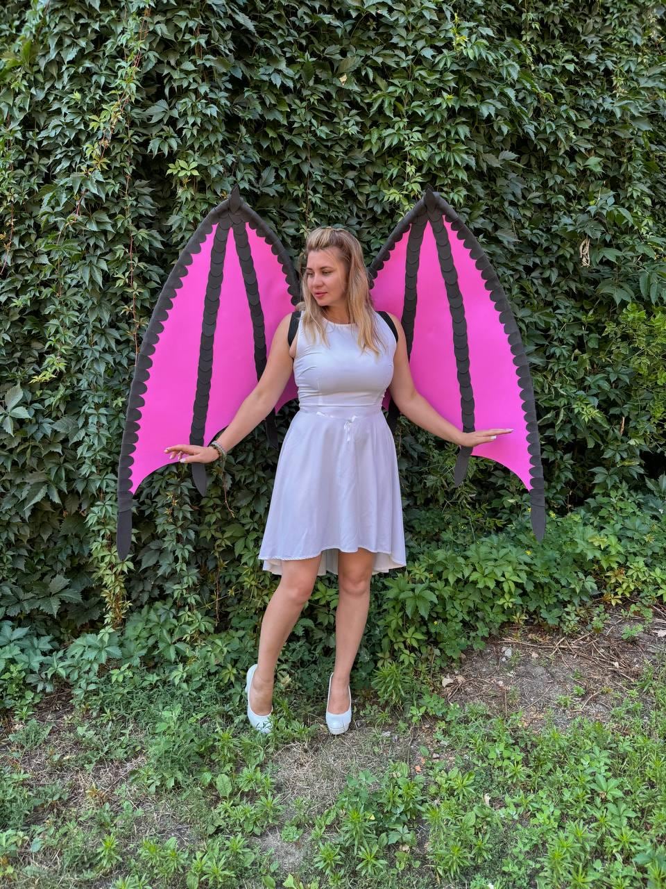 May include: A sleeveless white dress with a high-low hemline paired with large, detachable costume wings. The wings feature a vibrant pink interior with black structural ribs and a scalloped black border, secured by black shoulder straps.
