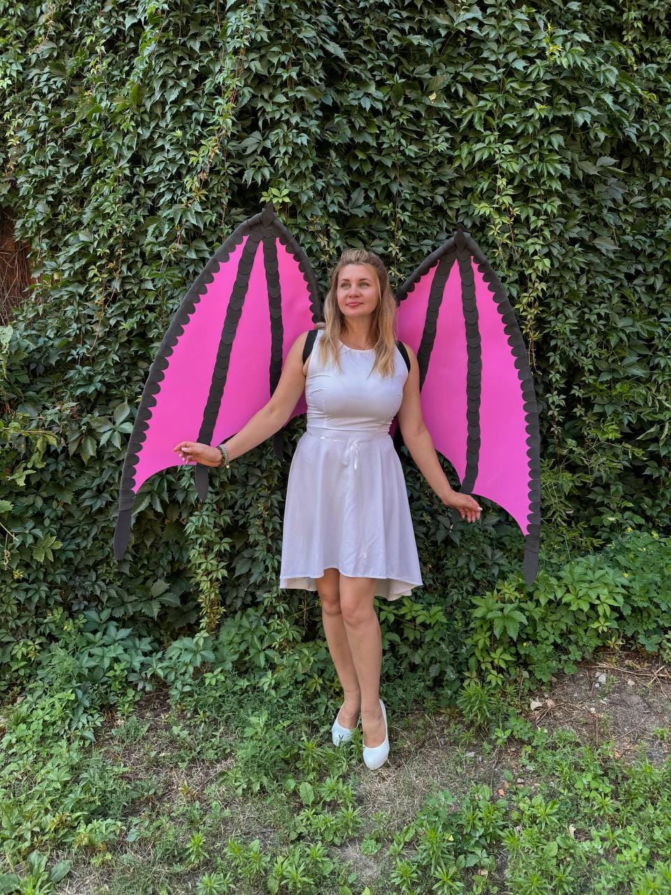 May include: A white sleeveless dress with a high neckline and a flared skirt, paired with large, detachable bat-style wings. The wings feature bright pink panels with black scalloped edges and black structural ribs, worn with black shoulder straps.