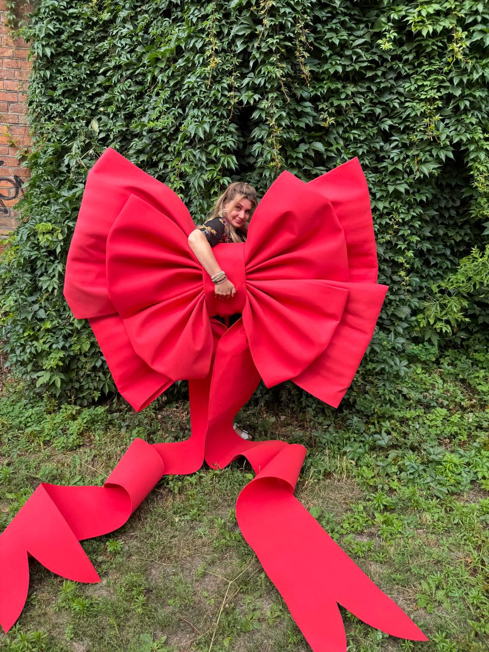 May include: An oversized, bright red decorative bow featuring large, voluminous loops and two long, trailing ribbon tails that extend downward. The bow is constructed with a smooth, matte finish and structured folds, creating a bold, prominent display.