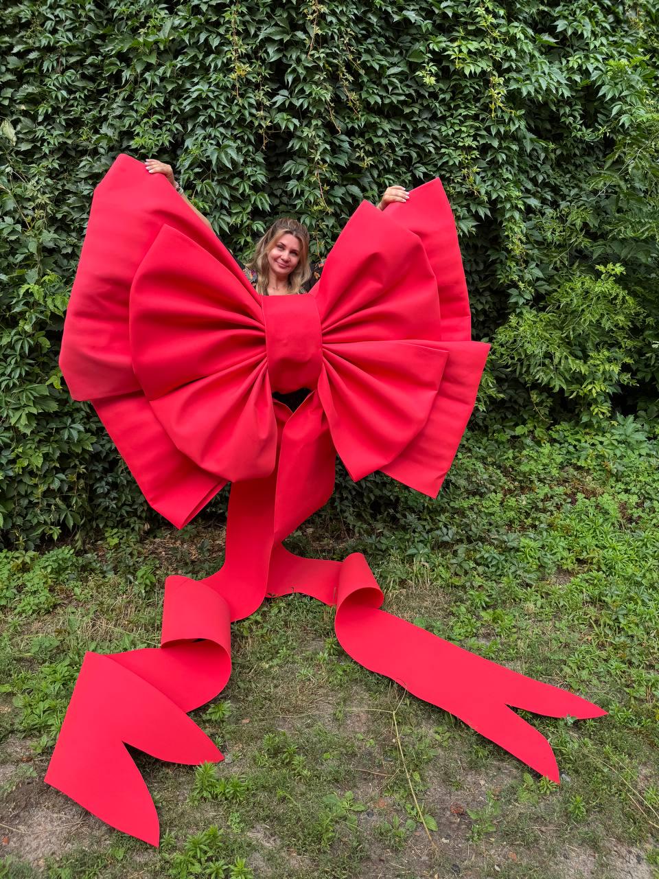 May include: An oversized, bright red decorative bow constructed with stiff, structured fabric. The bow features two large, voluminous loops and two long, trailing ribbon tails that extend downward, each ending in a V-shaped notch at the bottom.