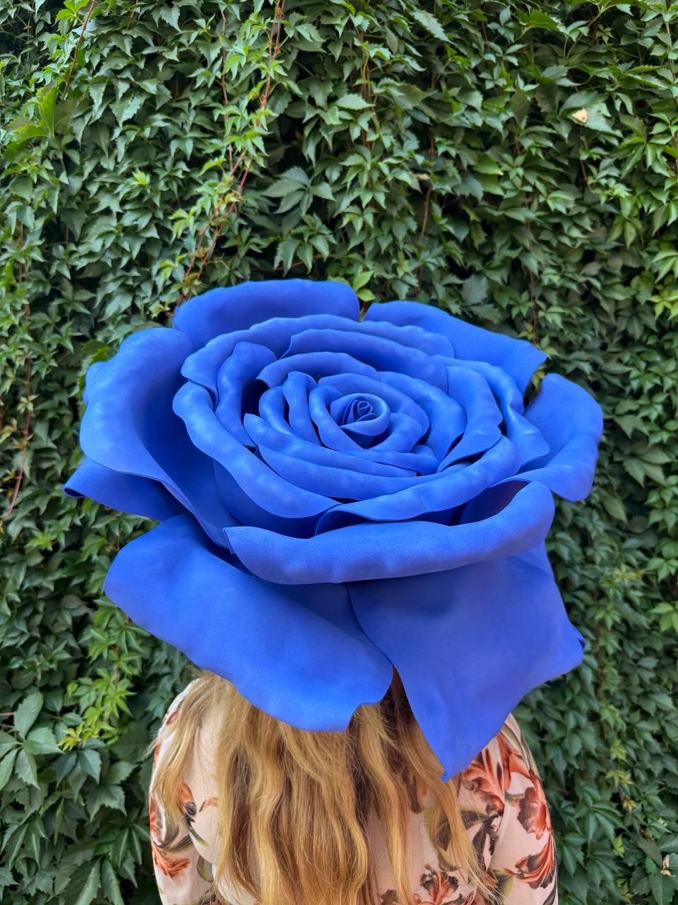 May include: A large, oversized artificial rose in a vibrant shade of blue. The flower features multiple layers of smooth, rounded petals that spiral inward toward a tightly closed center, creating a full, voluminous, and realistic floral shape.