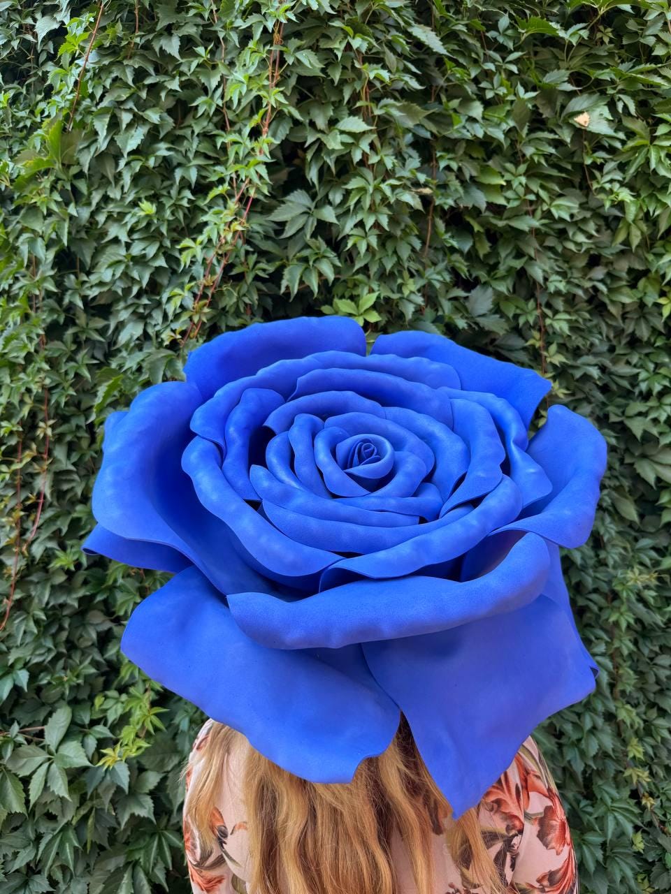 May include: A large, oversized artificial rose in a vibrant, solid blue color. The flower features multiple layers of wide, curved petals that form a dense, full bloom. The petals have a smooth, matte texture and are arranged in a circular pattern.