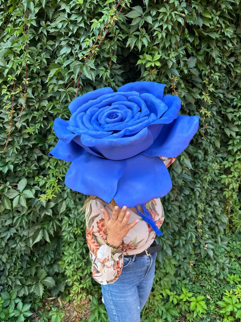 May include: A large, oversized artificial blue rose with layered, curved petals. The flower is held in front of the face, obscuring the head. The rose features a vibrant, solid blue color throughout its structure, creating a bold, sculptural appearance.