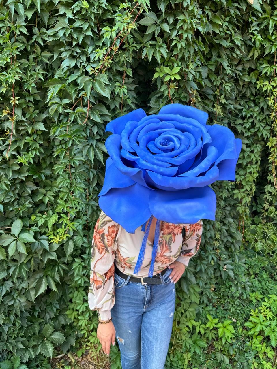 May include: A large, vibrant blue artificial rose head worn as a headpiece, paired with a long-sleeved beige blouse featuring a brown and orange floral pattern, and blue denim jeans with a black belt, set against a backdrop of green ivy leaves.