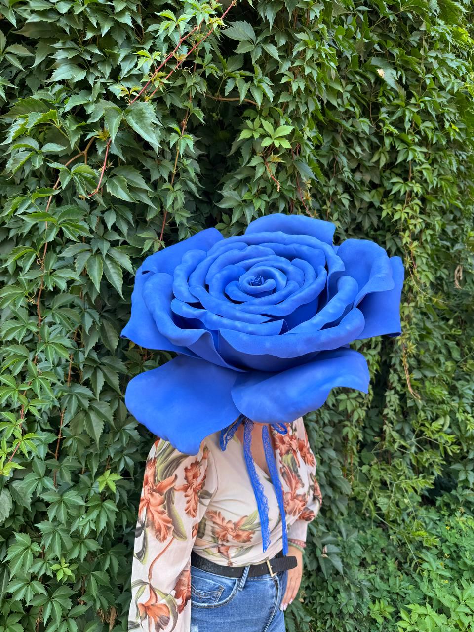May include: A large, oversized blue rose headpiece featuring layered, rounded petals. The flower is attached to a light-colored long-sleeved top with a floral print and blue jeans, secured with a thin blue ribbon tied under the chin.