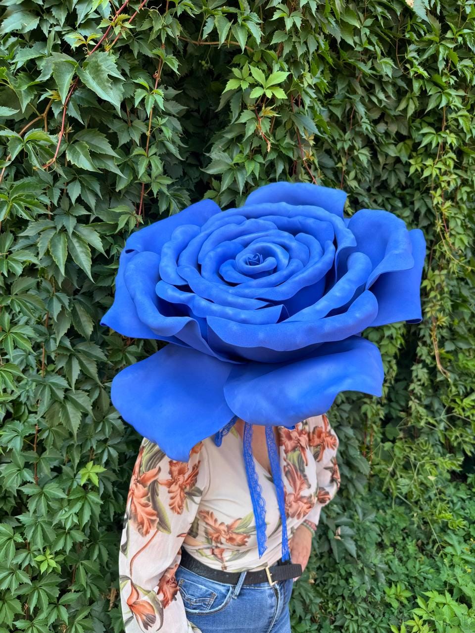 May include: A large, oversized blue rose headpiece featuring layered, curved petals in a vibrant matte blue finish. The rose is attached to a blue lace ribbon tie that hangs down, worn with a long-sleeved shirt with a floral print and blue jeans.