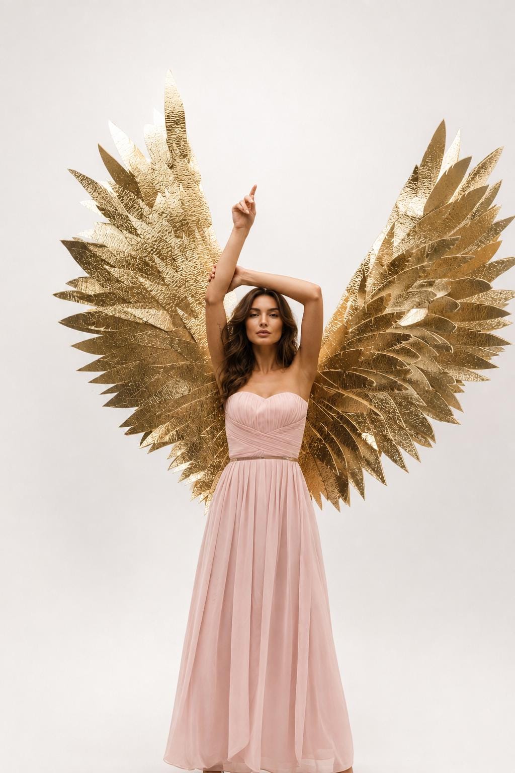 May include: A pair of large, textured gold wings with pointed feather-like segments, positioned behind a floor-length, strapless pale pink gown featuring a sweetheart neckline, a fitted bodice with pleated fabric, and a flowing skirt.