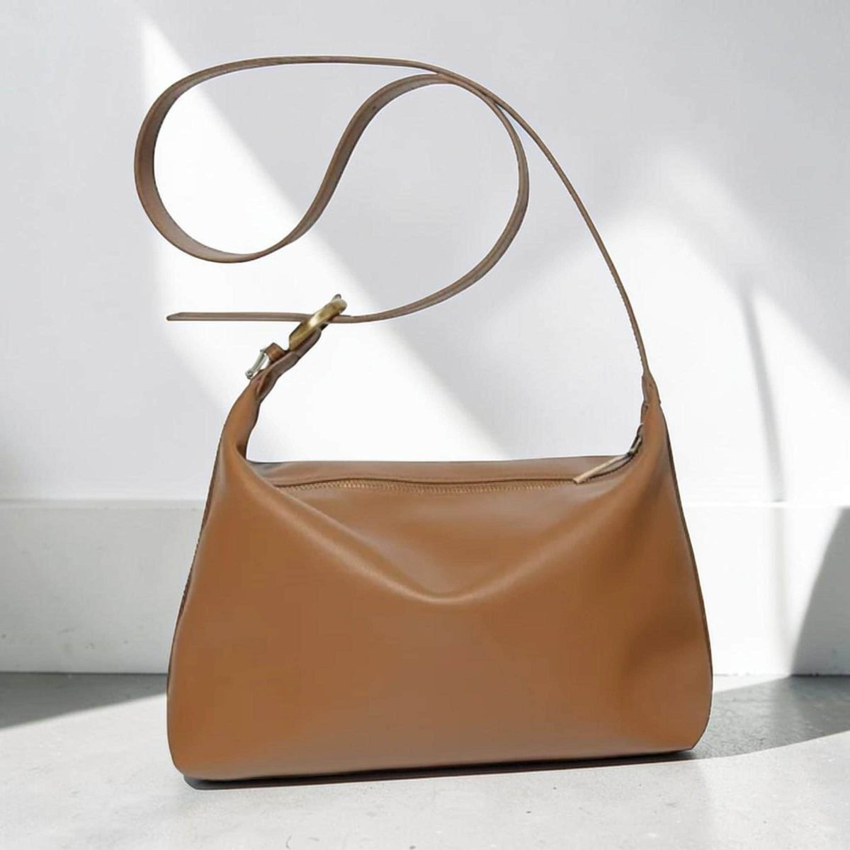 May include: A smooth, tan-colored shoulder bag with a rectangular silhouette and rounded corners. The bag features a long, adjustable strap with a metallic buckle and a top zipper closure, providing a clean and functional design for daily use.