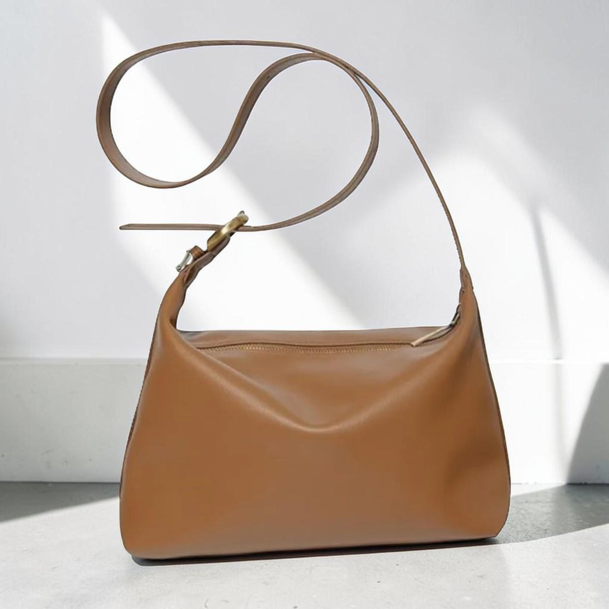May include: A smooth, tan-colored shoulder bag with a rectangular silhouette and rounded corners. The bag features a long, adjustable strap with a metallic buckle and a top zipper closure, providing a clean and functional design for daily use.