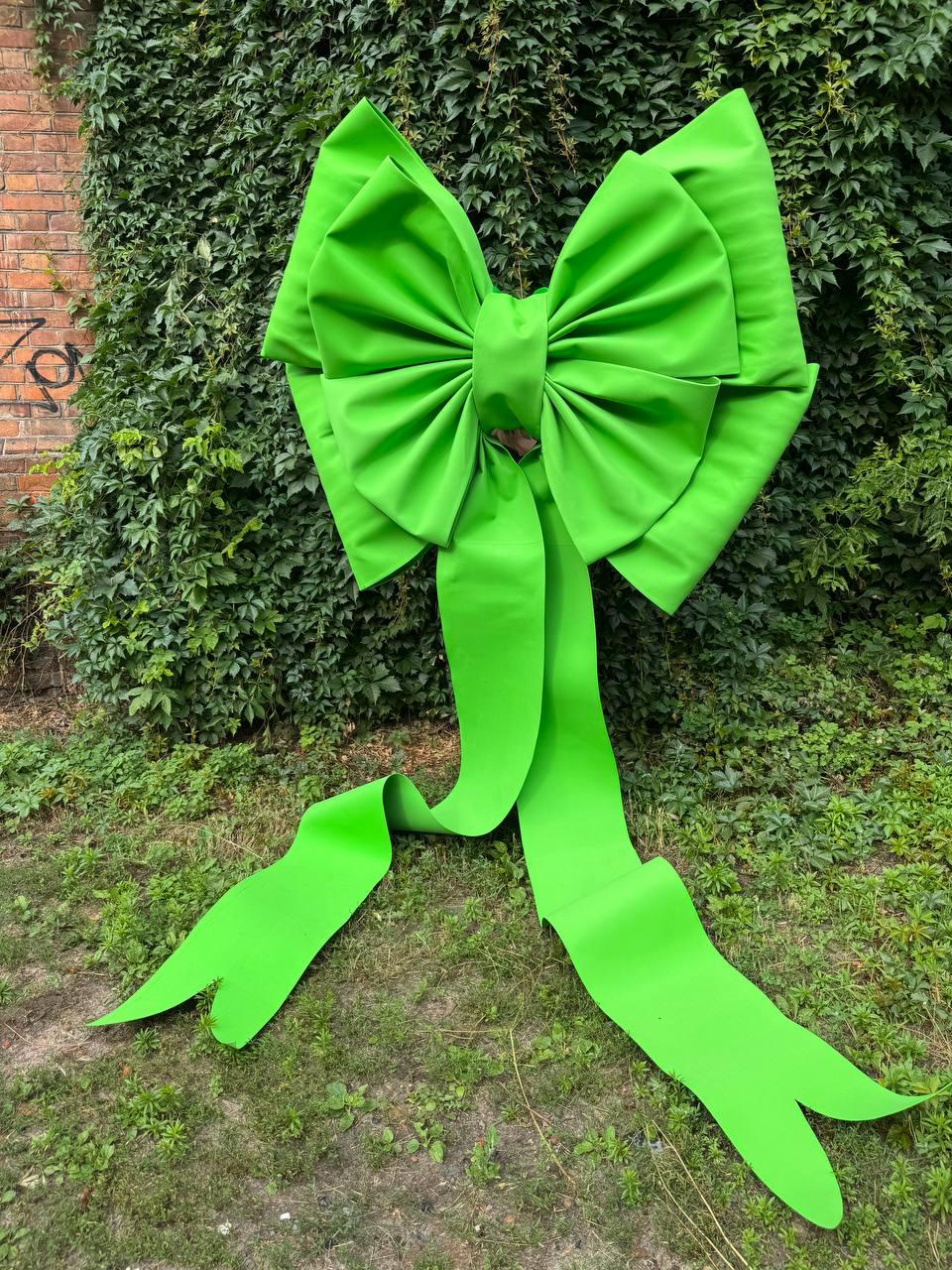 May include: A large, bright lime green decorative bow made of smooth, matte fabric. The bow features two wide, pleated loops and two long, flowing ribbon tails that extend downward, each ending in a distinct V-shaped notch at the bottom edge.