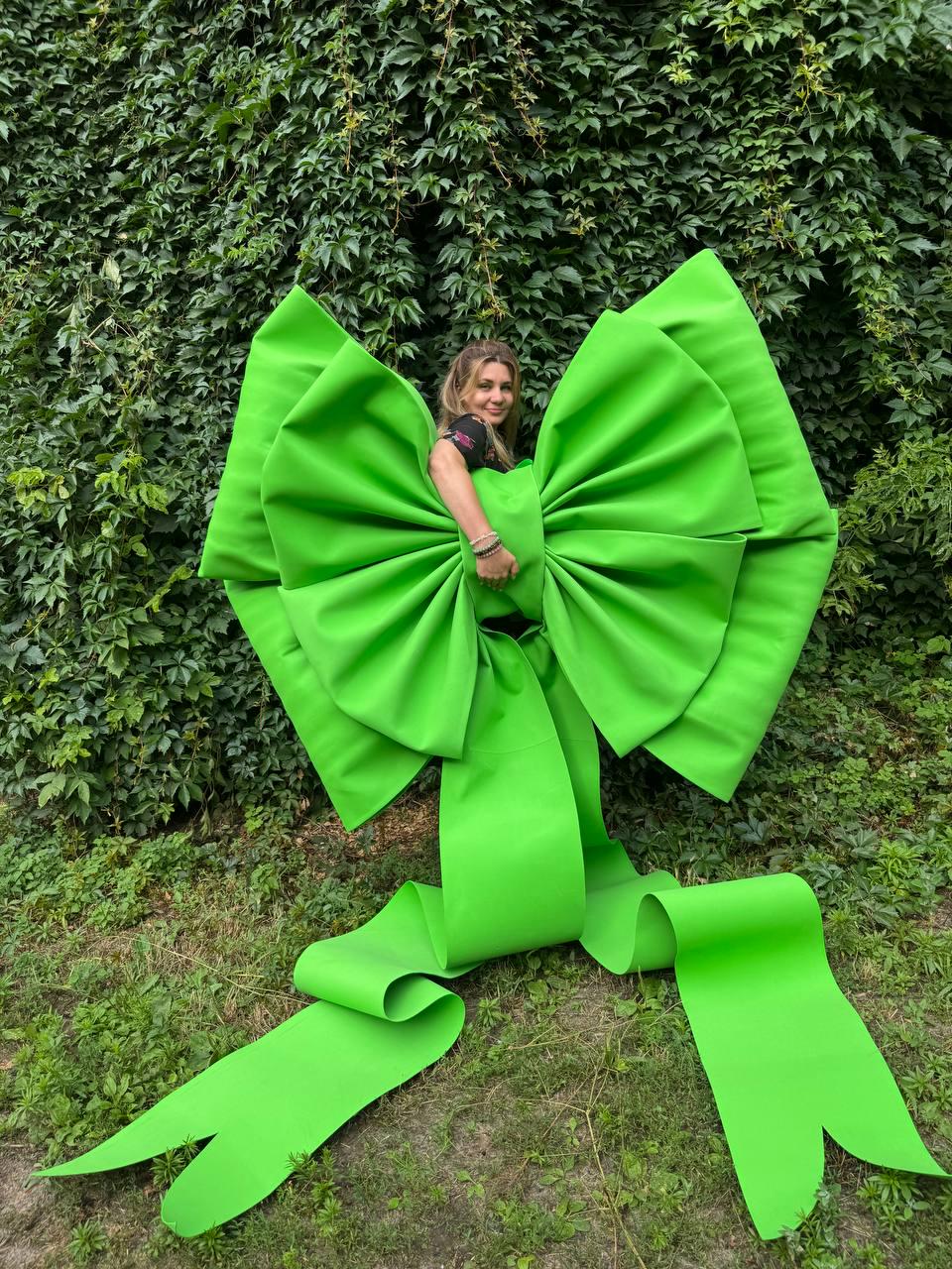 May include: A large, oversized bright lime green bow costume. The bow features wide, pleated fabric loops and two long, trailing ribbon ends that extend downward. The structure is designed to be worn, with a central knot section.
