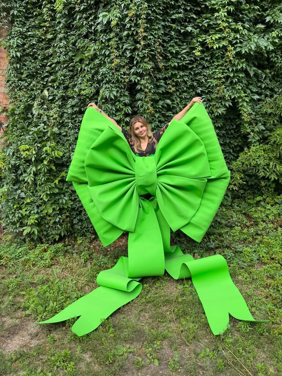 May include: An oversized, bright lime green decorative bow made of stiff material. The bow features large, voluminous loops and two long, wide ribbon tails that extend downward, creating a bold, structured shape with a smooth, matte finish.