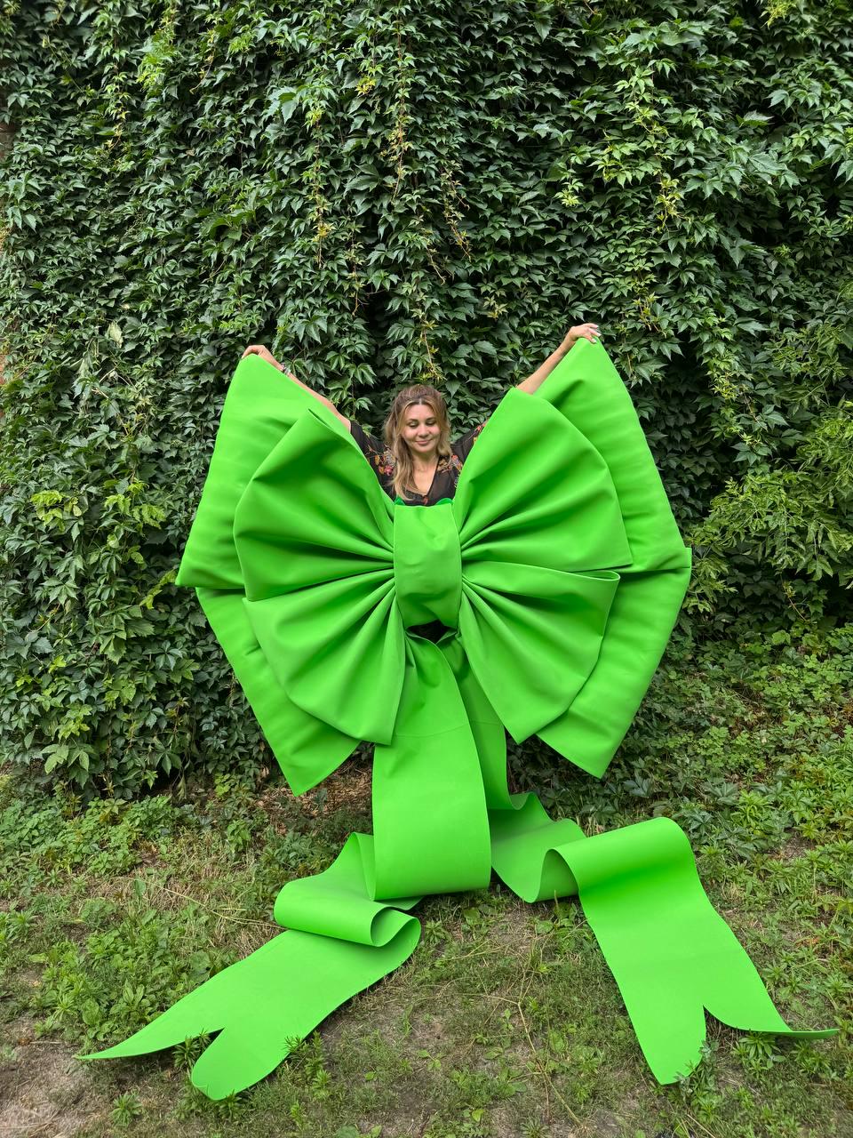 May include: An oversized, bright lime green decorative bow constructed from stiff material. The bow features two large, pleated loops and two long, wide ribbon tails that extend downward, creating a bold, sculptural accessory with a smooth finish.