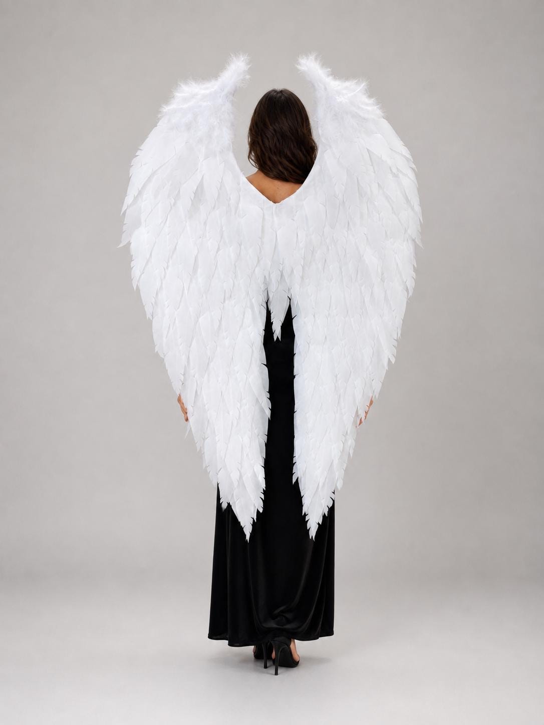 May include: Large white feathered angel wings designed to be worn on the back. The wings feature layered white feathers that extend upwards and outwards, creating a pointed shape. The wings are attached to a frame that rests against the back.